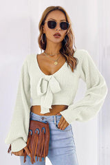 Woman wearing an off-white cropped tie-front sweater with long puffed sleeves and light wash jeans, holding a brown fringed handbag