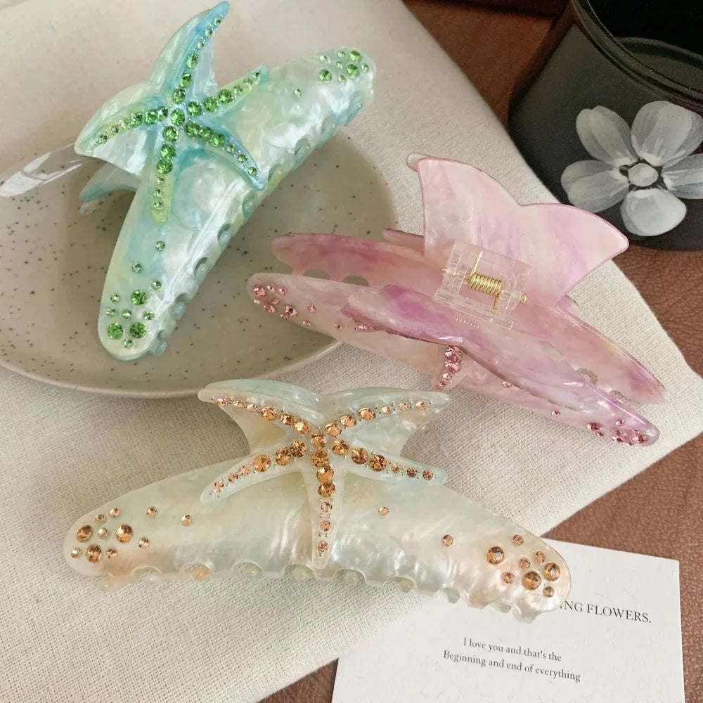 Three decorative hair claw clips arranged on a textured surface. One is blue with green rhinestones, one is pink with pink rhinestones, and one is white with gold rhinestones. All feature a starfish design.