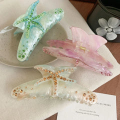 Three decorative hair claw clips arranged on a textured surface. One is blue with green rhinestones, one is pink with pink rhinestones, and one is white with gold rhinestones. All feature a starfish design.