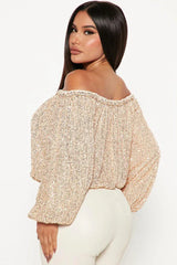 Woman wearing a champagne sequined off-the-shoulder top with balloon sleeves and white pants, back view