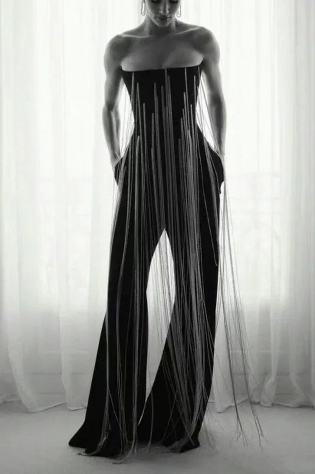 Model wearing a black strapless jumpsuit with long fringe detailing and wide legs, standing in front of a window.