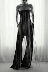 Model wearing a black strapless jumpsuit with long fringe detailing and wide legs, standing in front of a window.