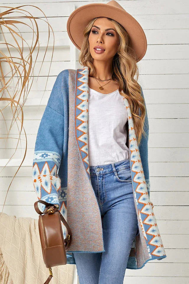 Woman wearing a long blue open-front cardigan with geometric print details, a white tank top, blue jeans, and a tan hat.