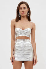 Front view of a woman with dark hair wearing a silver metallic ruched bustier top and matching ruched mini skirt, with star-shaped earrings.