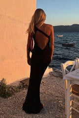 Woman from behind wearing a black sleeveless floor-length gown with a unique strap detail across the back, standing by the water at sunset.