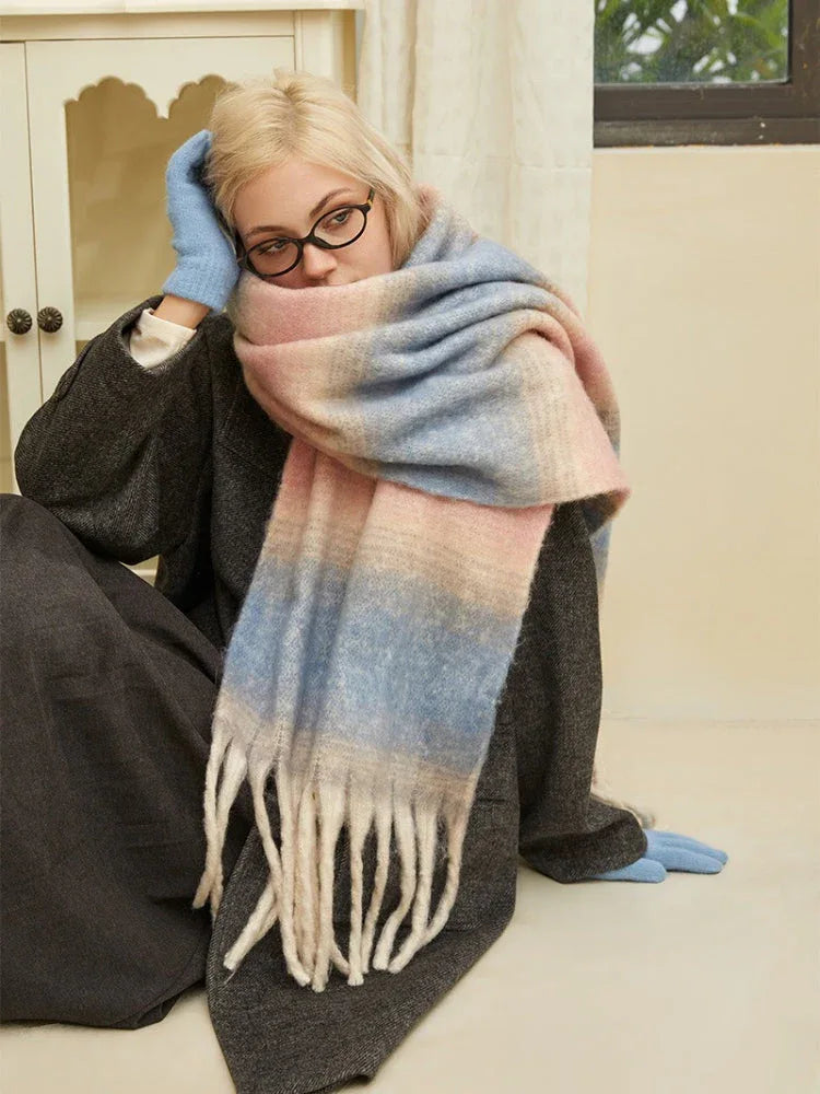 Woman sitting on the floor wearing a charcoal gray coat, light blue gloves, and a large ombre scarf with pink, blue, and cream colors. She is looking towards the camera.