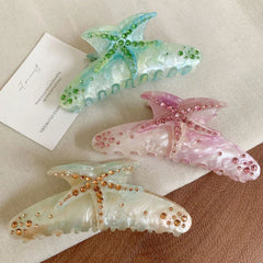 A top-down view of three stylish hair claw clips with starfish motifs and rhinestone accents, set against a light-colored fabric and a small card with text.