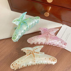 Three fashion hair claw clips, each designed with a starfish shape and decorated with vibrant rhinestones, are showcased on a surface with a wooden drawer in the background.