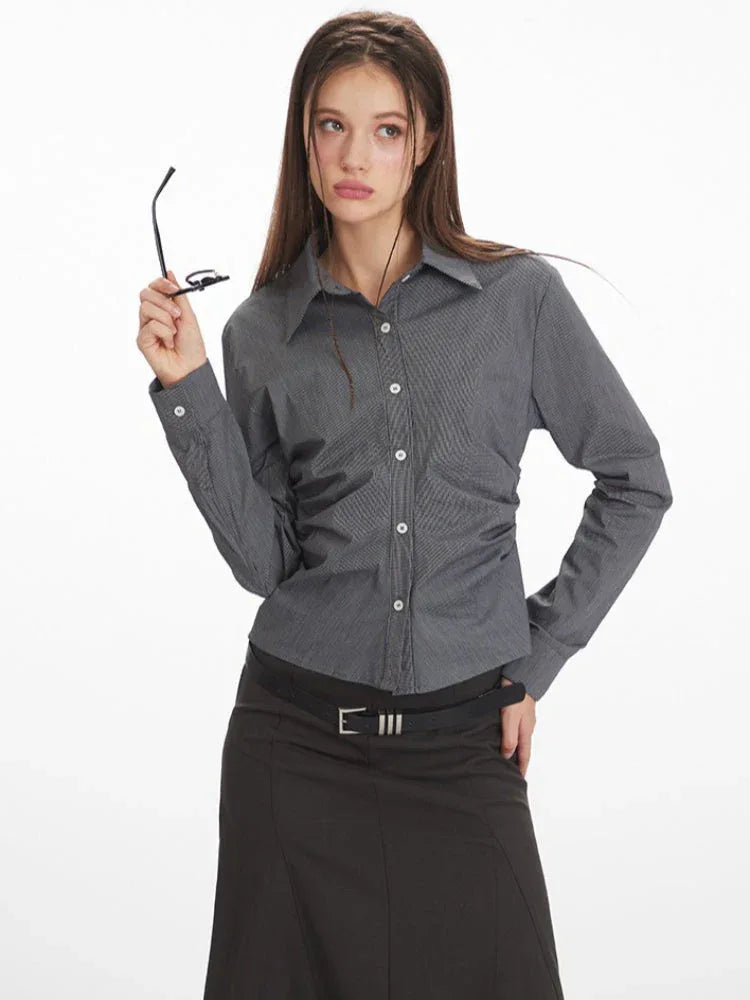 Woman wearing a grey long-sleeved button-down shirt with ruching detail, holding her glasses.