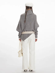 Woman wearing a grey asymmetrical knit poncho with a turtleneck, white pants, and a white belt, back view