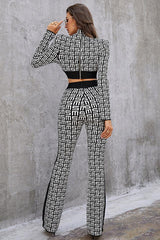 Back view of a woman in a black and white monogram print knit cropped long sleeve top with shoulder pads and matching flare pants with a contrasting side stripe, standing against a textured gray wall