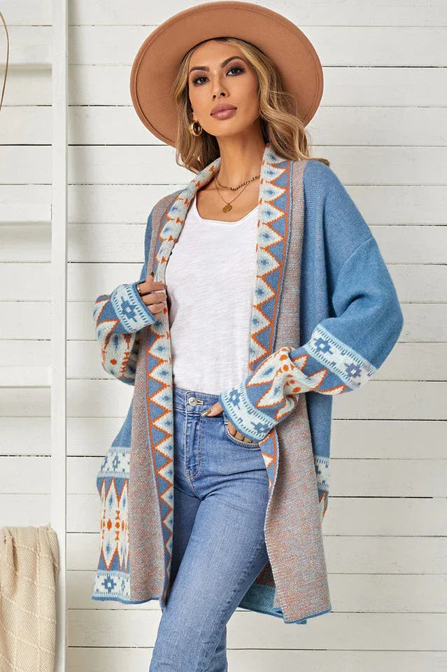 Woman wearing a long blue open-front cardigan with geometric print details, a white tank top, and blue jeans, holding the cardigan open.