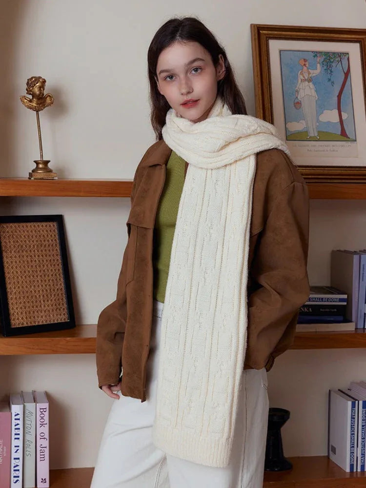 Woman standing in front of shelves, wearing a cream-colored cable knit scarf and brown jacket.