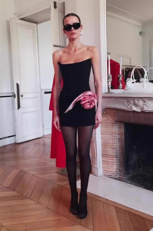 Model wearing a black strapless mini dress with a pink rose embellishment, black sheer tights, and sunglasses in an indoor setting.
