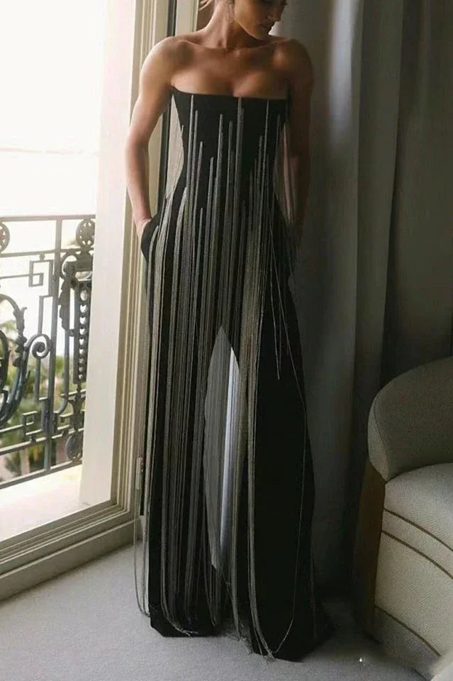 Full-length view of a model in a black strapless wide-leg jumpsuit with fringe detailing, posing near a window.