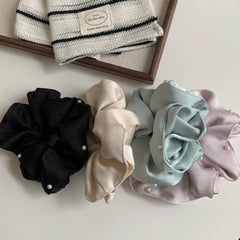 Four scrunchies in black, cream, light blue, and pastel pink, each with pearl accents, are displayed on a white surface next to a wooden tray containing a striped fabric item.