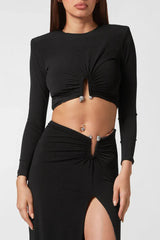 Close-up of a woman wearing a black long-sleeve crop top with a cut-out detail and a black maxi skirt with a high slit and embellished hardware.