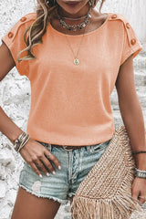 Woman wearing a peach short-sleeved t-shirt with button details on the shoulders and denim shorts.