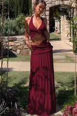Model wearing a burgundy two-piece set with a halter neck crop top and a tiered maxi skirt, posing outdoors in a garden setting
