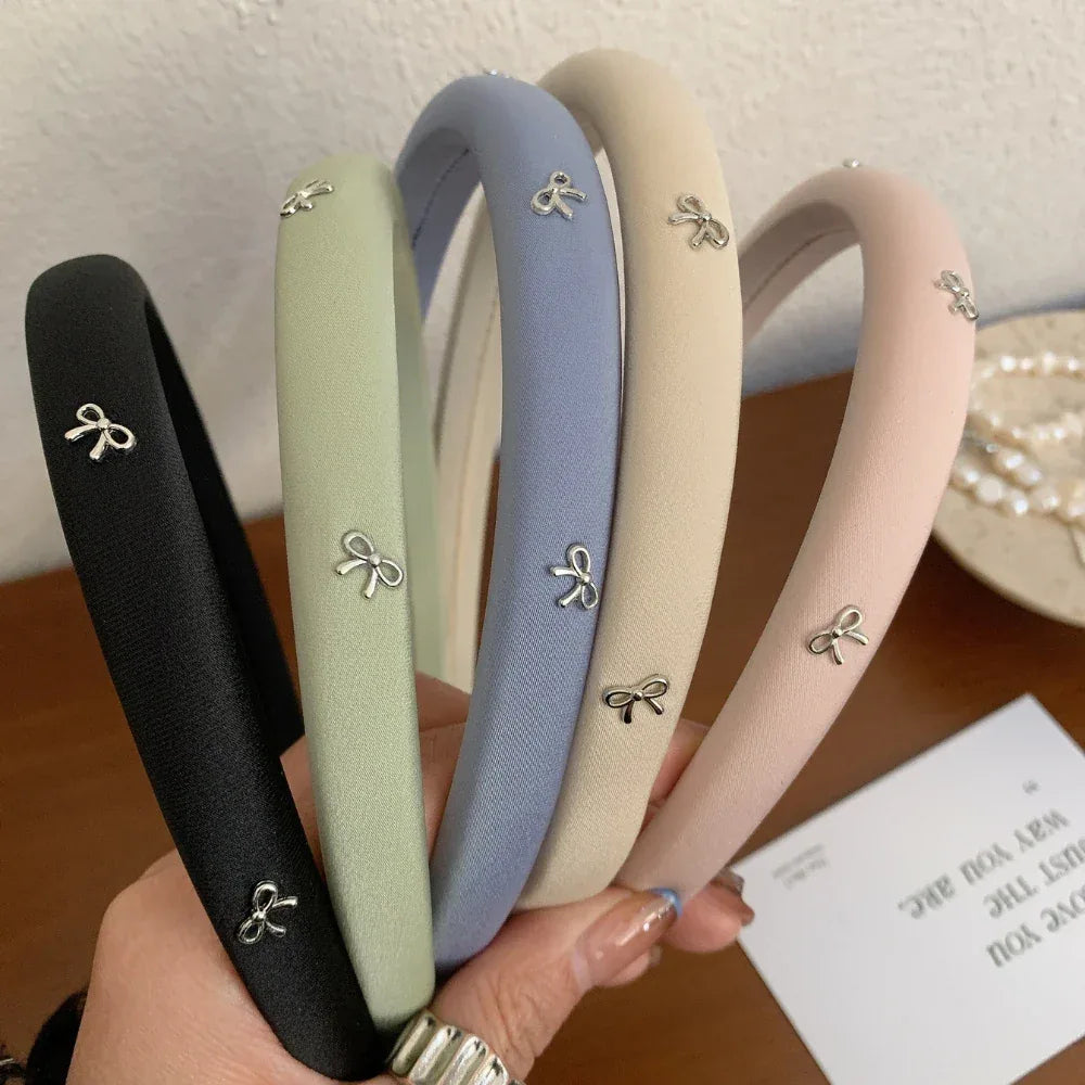 Collection of padded headbands in black, sage green, sky blue, beige, and blush pink, each adorned with small silver bow charms