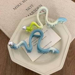 Close-up of light blue, green, and white S-shaped hair claw clips with pearls on a saucer