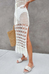 Close-up of the lower half of a woman's body wearing a white crochet knit maxi dress with a fringe hem and side slit, and sandals.