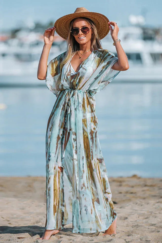 Full-length shot of a woman on a beach wearing a light, airy maxi dress with a watercolor print, a deep V-neck, and a straw hat.