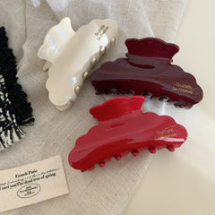 Three glossy hair claw clips in burgundy, red, and cream colors arranged on a textured fabric surface. Each clip features a cloud-like shape and embossed gold lettering.