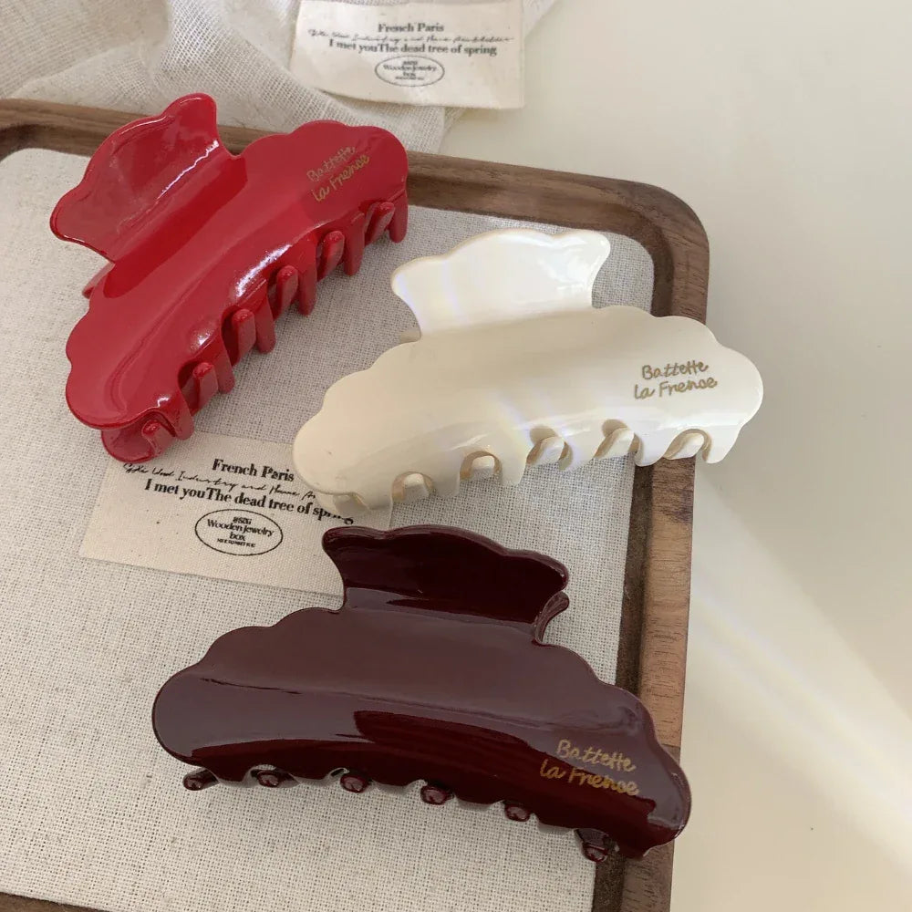 A flat lay arrangement of three glossy hair claw clips in red, cream, and burgundy, placed on a light-colored textured fabric. A small tag with French text is also visible.