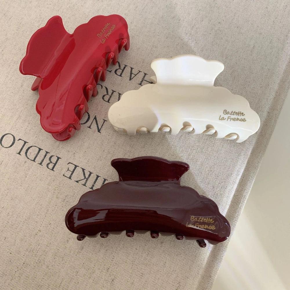 A group of three glossy hair claw clips in red, cream, and burgundy, displayed on a textured fabric surface. The clips have a distinct cloud shape and gold lettering.