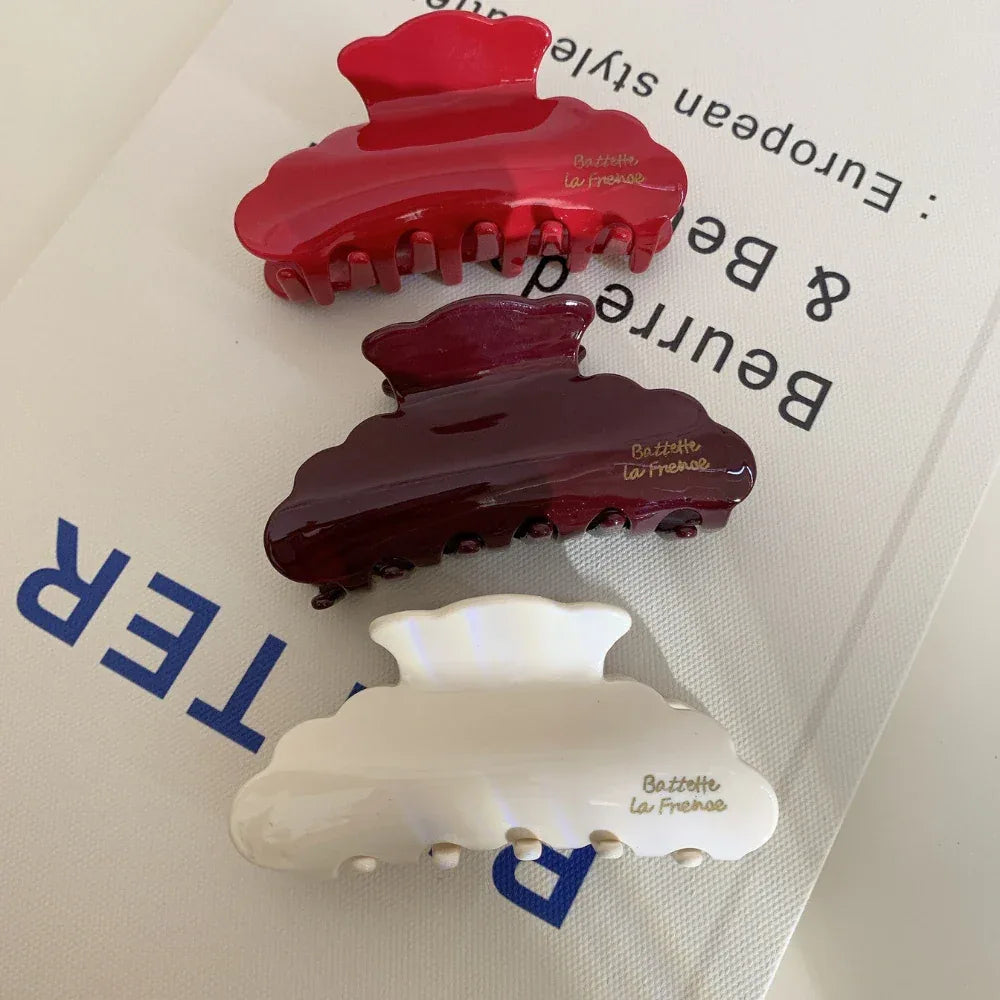 A top-down view of three glossy hair claw clips in red, burgundy, and cream, stacked on a surface with text. The clips are cloud-shaped with embossed gold lettering.
