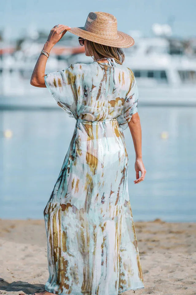 Rear view of a woman on a beach wearing a watercolor print maxi dress with flowing sleeves and a tie-waist detail.