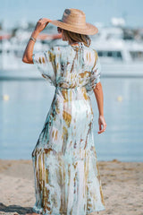 Rear view of a woman on a beach wearing a watercolor print maxi dress with flowing sleeves and a tie-waist detail.