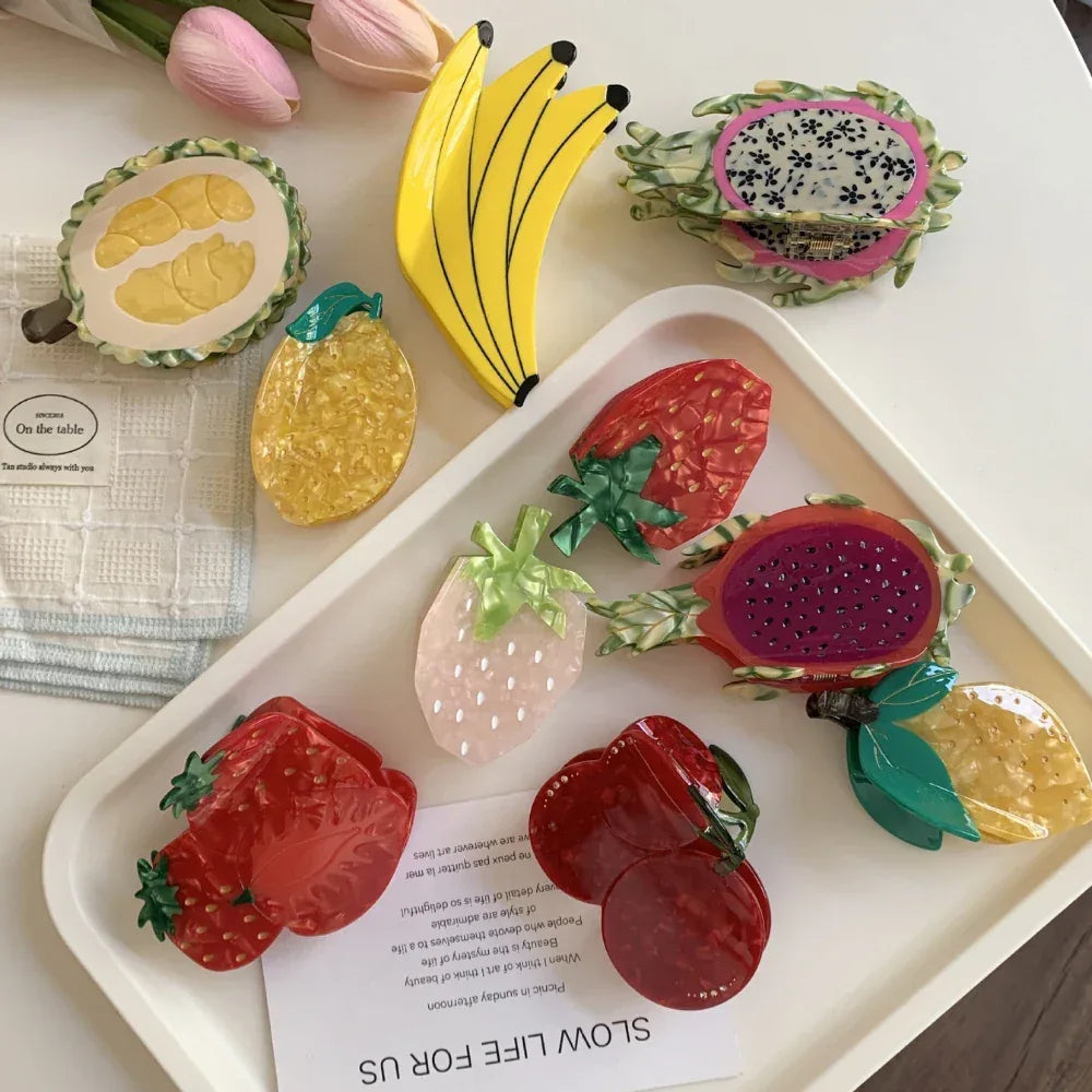 A collection of fruit-shaped hair clips arranged on a white tray, including bananas, strawberries, cherries, dragon fruit, durian, and lemon.