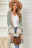 Woman wearing a long green open-front cardigan with geometric print details, a white tank top, blue jeans, and a tan hat.