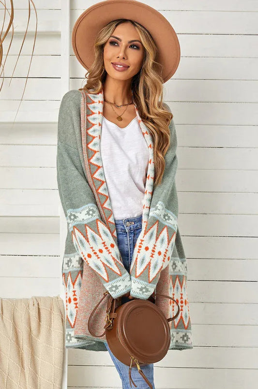 Woman wearing a long green open-front cardigan with geometric print details, a white tank top, blue jeans, and a tan hat.