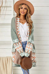 Woman wearing a long green open-front cardigan with geometric print details, a white tank top, blue jeans, and a tan hat.