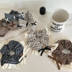 A variety of striped scrunchies with lace trim and bow accents are laid out on a white surface, alongside a mug and perfume bottle.