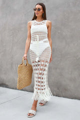 Woman wearing a white crochet knit maxi dress with a drawstring waist and fringe hem, holding a straw tote bag.