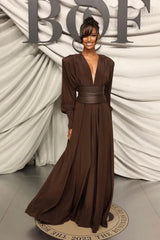 A woman in a brown draped maxi dress with a cinched waist and dramatic sleeves stands with her hand on her hip.