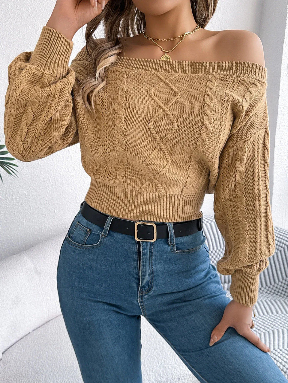 Woman wearing a tan off-the-shoulder cable knit cropped sweater with blue jeans and a black belt.