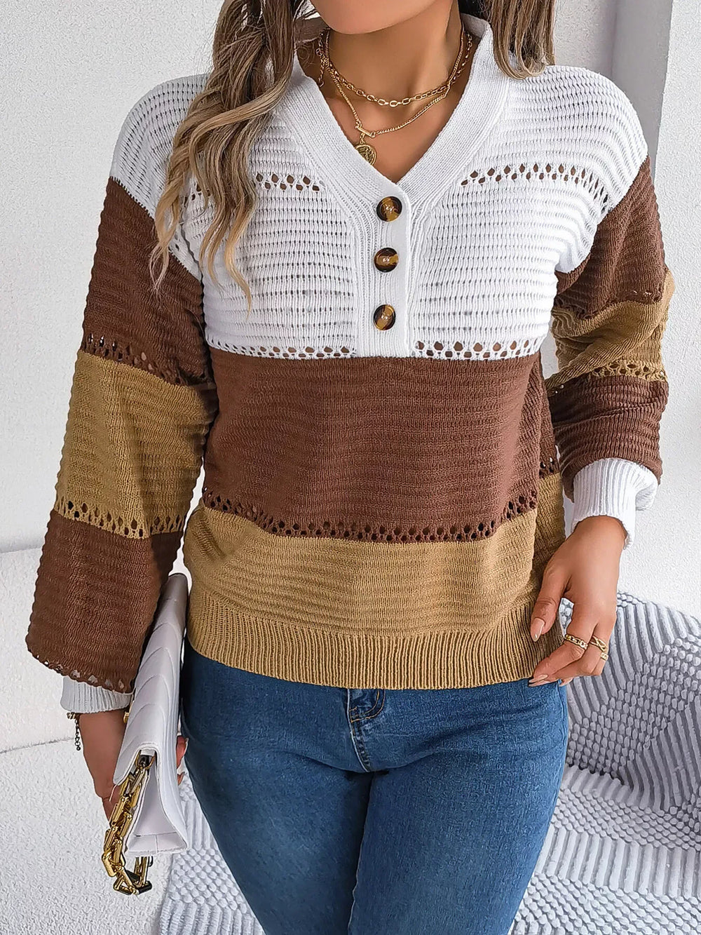 Woman wearing a brown, tan, and white color block knit sweater with a v-neck and button details, paired with blue jeans and holding a white clutch.