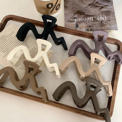 A collection of wavy claw clips in various colors including black, white, purple, beige, and brown, arranged on a wooden tray.