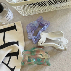 overhead view of three abstract figure hair claw clips in purple, white, and marbled green/pink/brown on a textured surface with a bottle of perfume and a white basket
