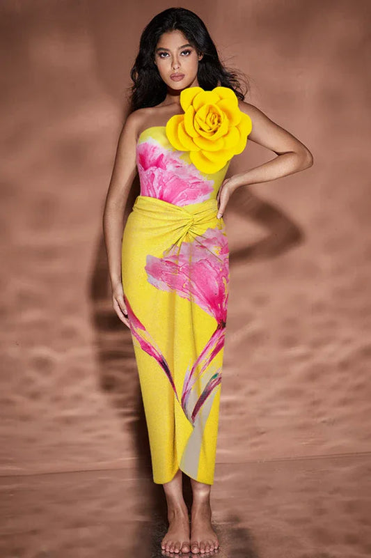 Woman in yellow one-piece swimsuit with floral print and large yellow flower embellishment on shoulder, standing with hand on hip.