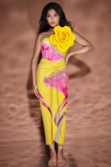 Woman in yellow one-piece swimsuit with floral print and large yellow flower embellishment on shoulder, standing with hand on hip.