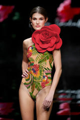 Fashion show runway image of a model wearing a one-piece swimsuit with a tropical parrot and palm leaf print, and a large red floral applique on the shoulder.