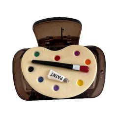A close-up product shot of a single brown translucent hair claw clip adorned with a heart-shaped art palette featuring colorful paint dots, a paintbrush, and a small tube labeled "PAINT."
