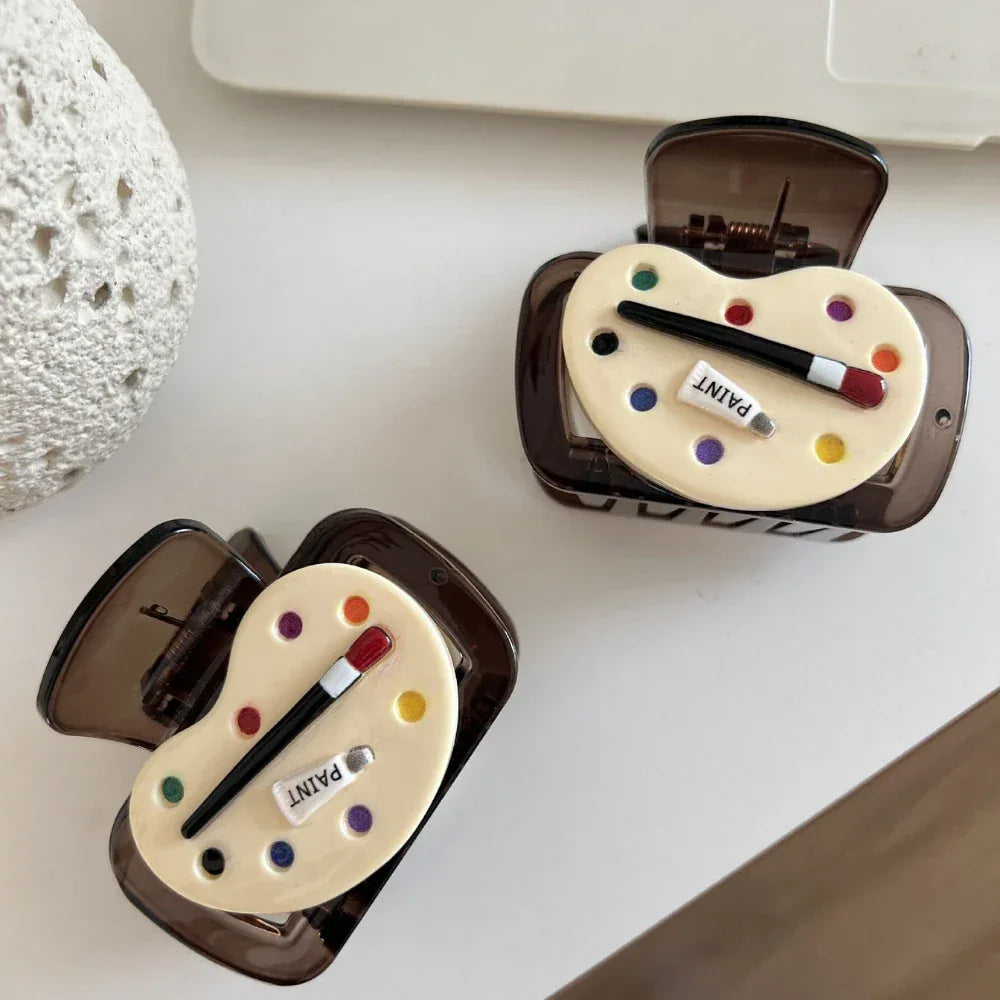 Two brown translucent hair claw clips, each decorated with an art palette design, are displayed on a white surface next to a textured white object and a laptop.