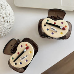 Two brown translucent hair claw clips, each decorated with an art palette design, are displayed on a white surface next to a textured white object and a laptop.